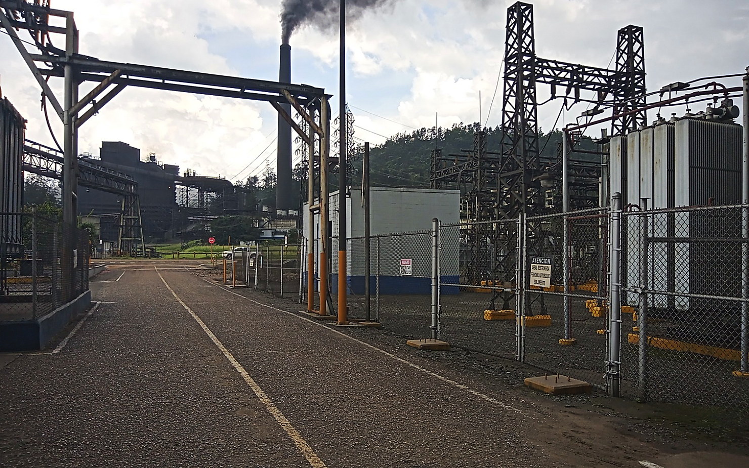 Patio de subestación con transformadores y pórticos junto a una planta industrial en operación
